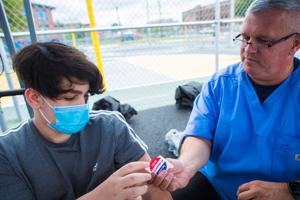  John Pizzo, a public health nurse for the Rhode Island National Guard hands a sticker to Victor Urizar, 15, of Providence after giving him a COVID-19 vaccine at Bucklin Park in August.