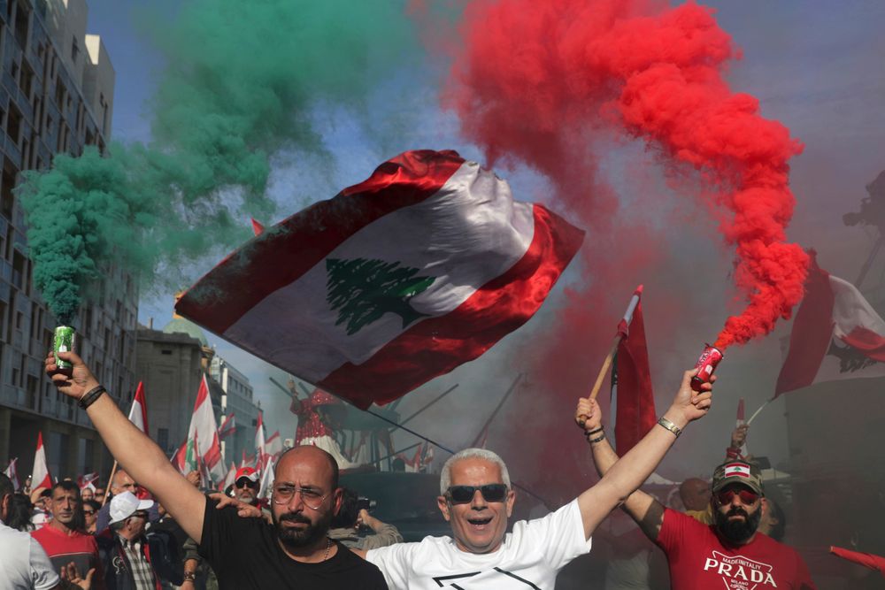 Anti-government protesters wave Lebanese national flags as hold smoke flares during separate civil parade at the Martyr square, in downtown Beirut, Lebanon, Friday, Nov. 22, 2019. Protesters gathered for alternative independence celebrations, converging by early afternoon on Martyrs’ Square in central Beirut, which used to be the traditional location of the official parade. Protesters have occupied the area, closing it off to traffic since mid-October. (AP Photo/Hassan Ammar) Anti-government protesters wave Lebanese national flags as hold smoke flares during separate civil parade at the Martyr square, in downtown Beirut, Lebanon, Friday, Nov. 22, 2019. Protesters gathered for alternative independence celebrations, converging by early afternoon on Martyrs’ Square in central Beirut, which used to be the traditional location of the official parade. Protesters have occupied the area, closing it off to traffic since mid-October. (AP Photo/Hassan Ammar)