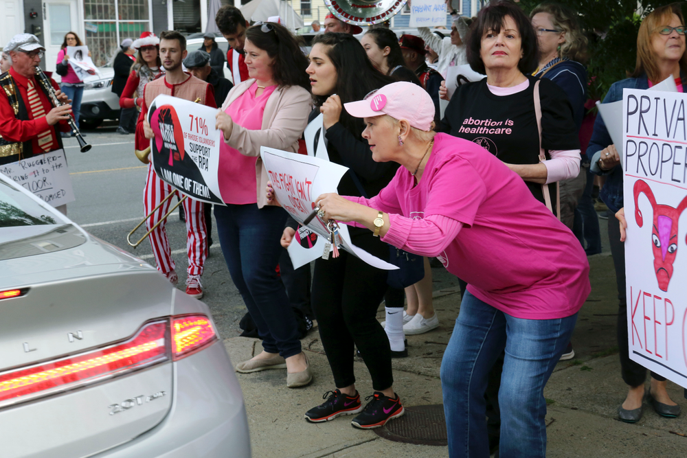 In this Thursday, May 23, 2019, photo, Nancy St Germain, 71, right, of Warwick, R.I., leans forward to show her sign to a driver leaving a fundraiser for the Rhode Island Senate Democrats political action committee in Providence, R.I. Her sign reads, In this Thursday, May 23, 2019, photo, Nancy St Germain, 71, right, of Warwick, R.I., leans forward to show her sign to a driver leaving a fundraiser for the Rhode Island Senate Democrats political action committee in Providence, R.I. Her sign reads,