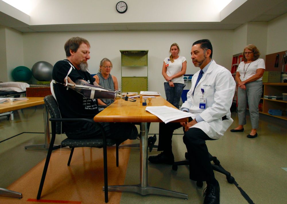 In this Aug. 19, 2019 photo, Greg Manteufel tries out a new prosthetic arm during occupational therapy at Froedtert & the Medical College of Wisconsin in Milwaukee, as he talks to Dr. David Del Toro, the medical director for inpatient rehab. Manteufel lost parts of his arms and legs, as well as the skin of his nose and part of his upper lip from capnocytophaga, a bacteria commonly found in the saliva or cats and dogs that almost never leads to people getting sick, unless the person has a compromised immune system. He was perfectly healthy when he got sick in June of 2018. Over the last seven years, a team of researchers at Brigham and Women's Hospital in Boston, connected to Harvard Medical School, have tested other healthy people who were affected and developed a theory on why they were affected- a gene change in all the victims. (AP Photo/Carrie Antlfinger) In this Aug. 19, 2019 photo, Greg Manteufel tries out a new prosthetic arm during occupational therapy at Froedtert & the Medical College of Wisconsin in Milwaukee, as he talks to Dr. David Del Toro, the medical director for inpatient rehab. Manteufel lost parts of his arms and legs, as well as the skin of his nose and part of his upper lip from capnocytophaga, a bacteria commonly found in the saliva or cats and dogs that almost never leads to people getting sick, unless the person has a compromised immune system. He was perfectly healthy when he got sick in June of 2018. Over the last seven years, a team of researchers at Brigham and Women's Hospital in Boston, connected to Harvard Medical School, have tested other healthy people who were affected and developed a theory on why they were affected- a gene change in all the victims. (AP Photo/Carrie Antlfinger)